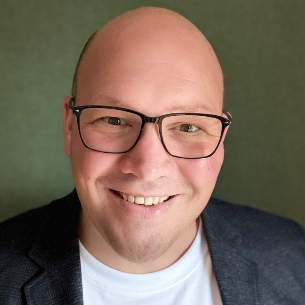 Mann mit Glatze, Brille und lächelndem Gesicht vor grünem Hintergrund.